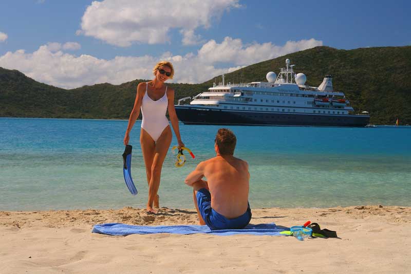 Photo d'une jeune femme en maillot de bain une pièce qui sort de la mer avec des palmes à la main, on voit un bateau de 
											croisière pour célibataires en arrière plan