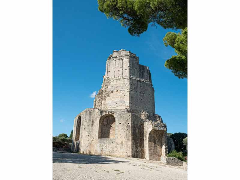 Vue de la tour Magne à Nîmes, vestige d’une tour romaine en pierre surplombant la ville, entourée de pins sous un ciel bleu clair