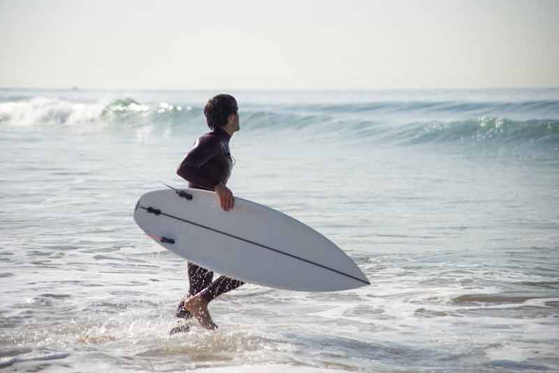 Surfeur entrant dans l’océan à Hourtin avec sa planche de surf, marchant dans l’eau face aux vagues sous une lumière douce
