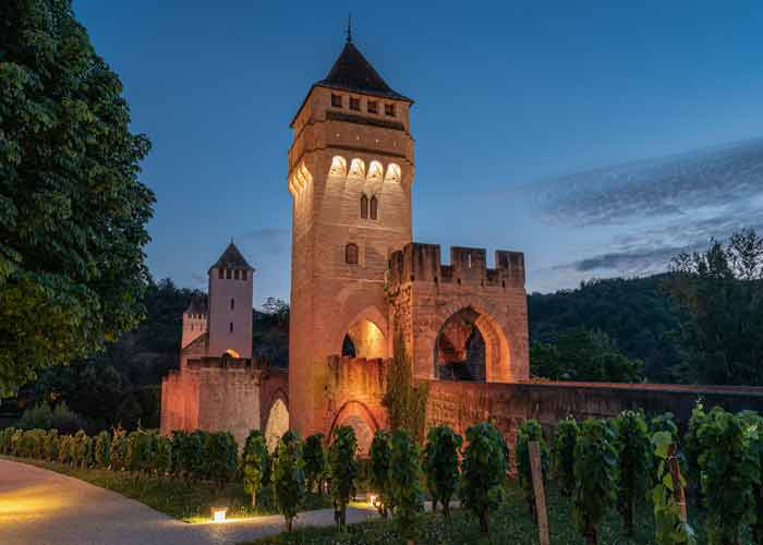 Pont Valentré à Cahors illuminé au crépuscule, avec ses tours médiévales en pierre et un sentier bordé de vignes au premier plan