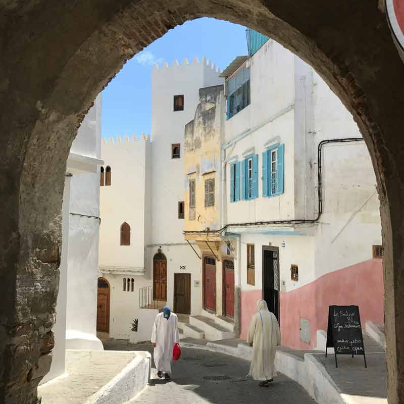 Vieille ruelle pittoresque de la médina de Tanger, encadrée par une arche en pierre, avec des façades blanchies et des volets 
							bleus