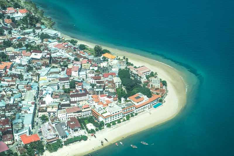 Vue aérienne de Stone Town à Zanzibar, avec ses bâtiments aux toits colorés bordés par une plage de sable blanc et l'océan Indien aux eaux turquoise