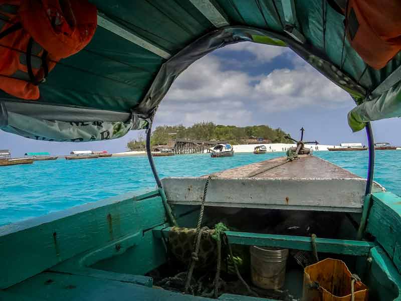 Vue depuis un bateau traditionnel en bois approchant une plage paradisiaque à Zanzibar, avec des eaux turquoise éclatantes et des embarcations ancrées près du rivage